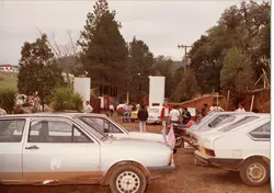 Uma fotografia colorida de época, provavelmente das décadas de 1980 ou 1990, mostrando um grupo de pessoas reunidas em uma área externa próxima a veículos estacionados. O cenário sugere um ambiente rural ou semi-urbano com vegetação e construções simples, comum em pequenas cidades brasileiras.