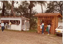 Uma fotografia colorida que mostra duas pequenas estruturas de madeira, possivelmente quiosques de venda de alimentos, com pessoas sendo atendidas. O ambiente sugere uma área externa ou de lazer, com vegetação ao fundo e um veículo estacionado em primeiro plano, remetendo ao estilo de vida e comércio informal de pequenas cidades brasileiras nas décadas de 1980 ou 1990.