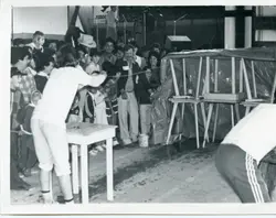 Uma fotografia em preto e branco que registra um evento comunitário ou recreativo, possivelmente em um ginásio ou pavilhão coberto. Vê-se um grupo de pessoas, incluindo crianças e adultos, observando uma atividade que envolve mesas e uma estrutura coberta por lona, sugerindo uma gincana ou feira local típica de meados da década de 1980.