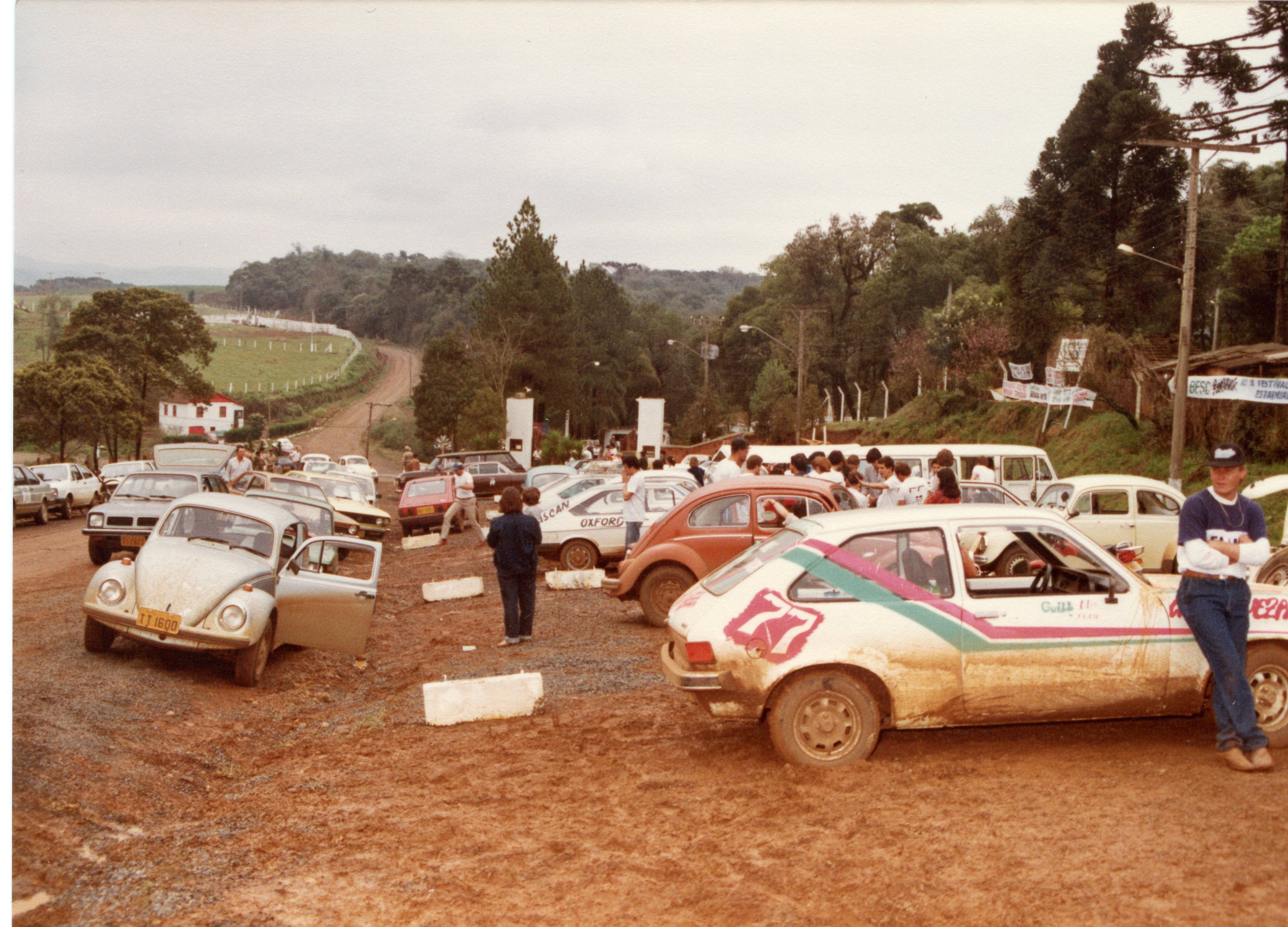 Uma cena de um evento de automobilismo, possivelmente um rali, realizado em um terreno de terra batida com muitos carros clássicos brasileiros, como Fuscas e Chevettes, cobertos de lama. O ambiente sugere uma área rural ou periurbana, com espectadores e participantes reunidos em torno dos veículos, indicando uma competição amadora ou regional típica das décadas de 1980 ou 1990.