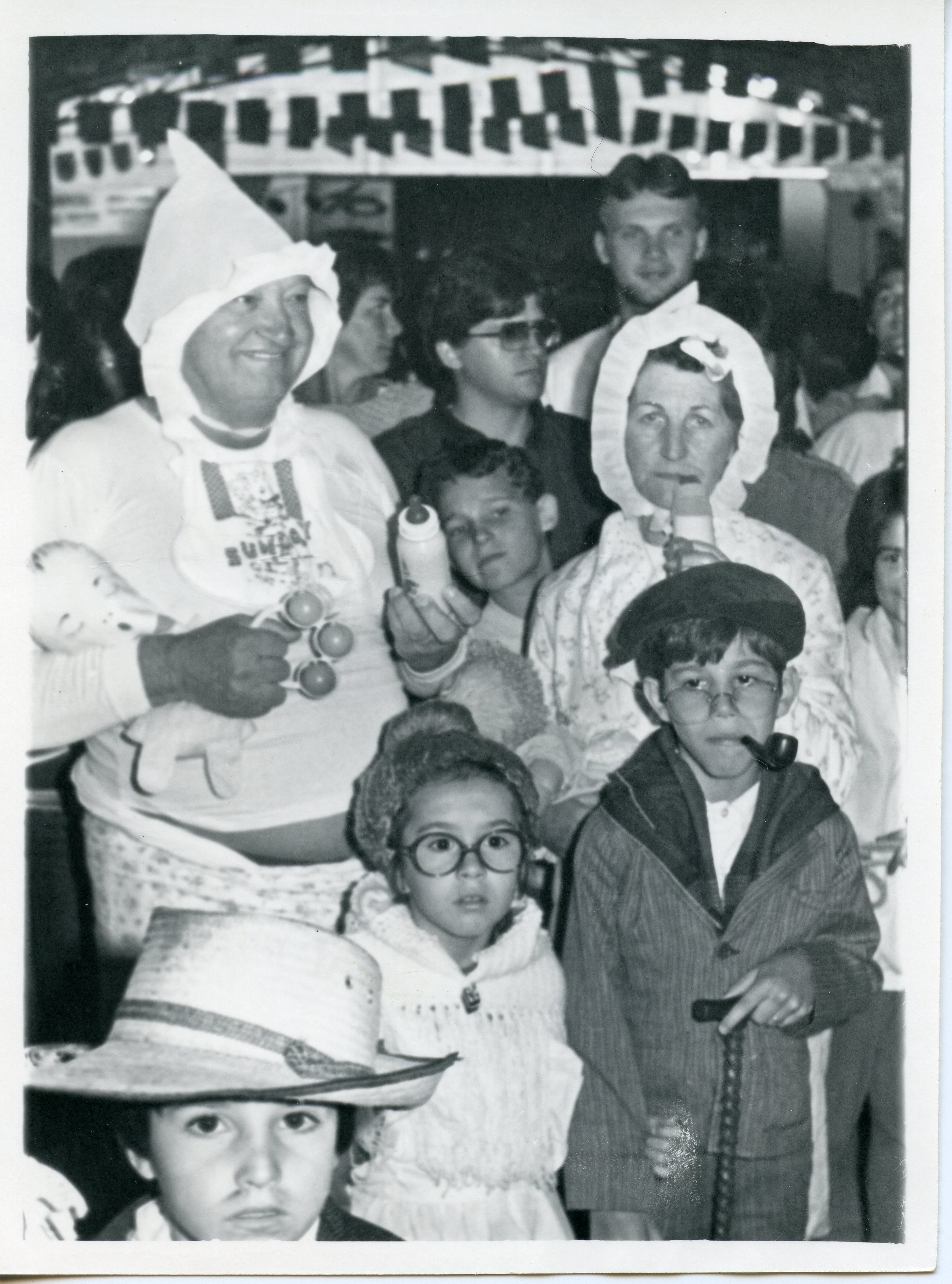 Uma fotografia em preto e branco capturando um grupo de pessoas fantasiadas, possivelmente em um evento comunitário ou festa temática. Os participantes vestem roupas que remetem a bebês e idosos, sugerindo uma brincadeira ou desfile, com elementos como mamadeiras, gorros e óculos, típicos de celebrações escolares ou sociais brasileiras das décadas de 1980 ou 1990.