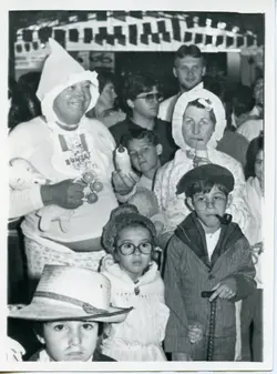 Uma fotografia em preto e branco capturando um grupo de pessoas fantasiadas, possivelmente em um evento comunitário ou festa temática. Os participantes vestem roupas que remetem a bebês e idosos, sugerindo uma brincadeira ou desfile, com elementos como mamadeiras, gorros e óculos, típicos de celebrações escolares ou sociais brasileiras das décadas de 1980 ou 1990.
