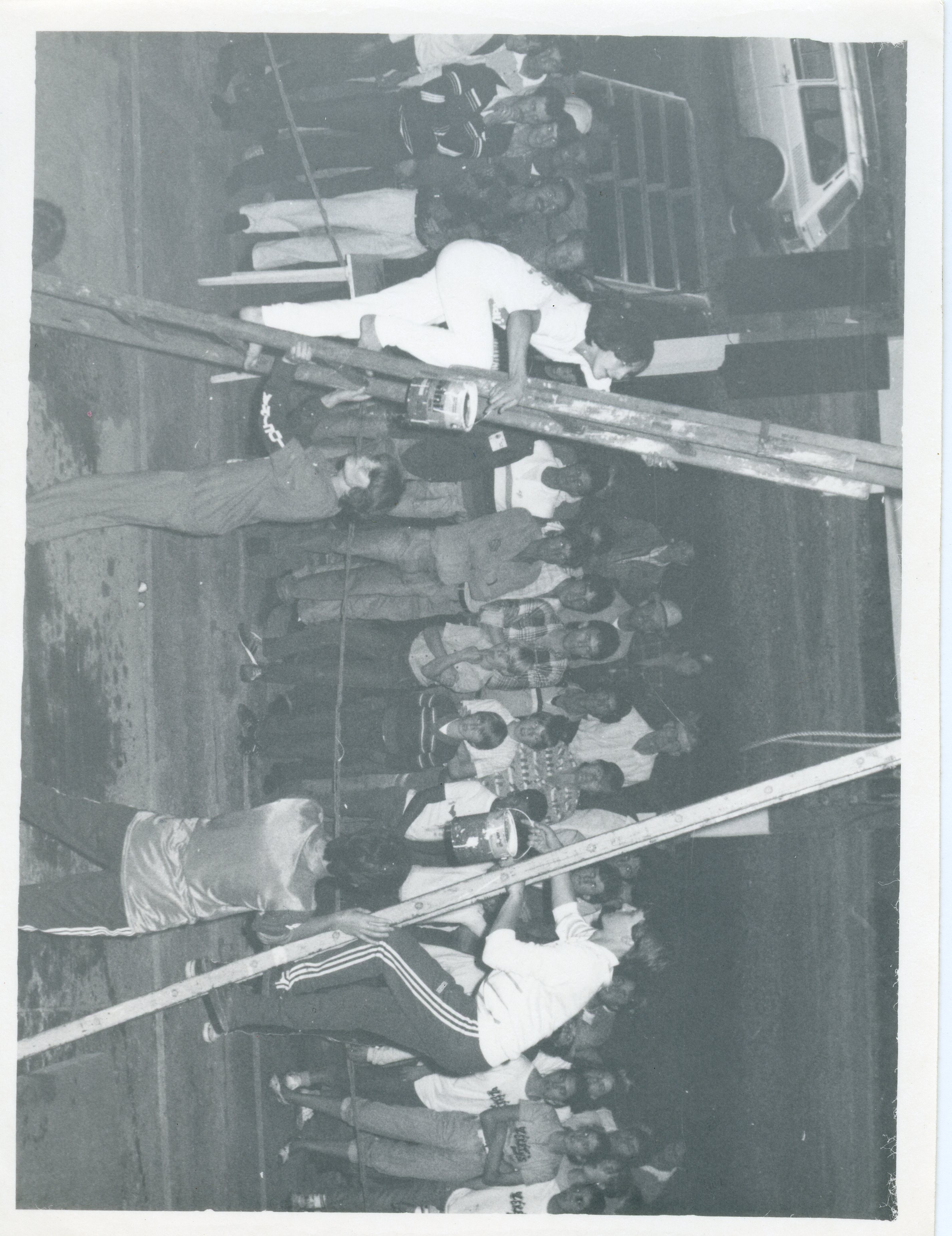 Fotografia em preto e branco que registra um evento comunitário ou esportivo ao ar livre, possivelmente uma gincana ou competição recreativa. Várias pessoas estão reunidas, com alguns participantes escalando estruturas de madeira ou andaimes, sugerindo uma atividade de lazer ou desafio físico típica de meados da década de 1980.