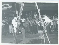 Fotografia em preto e branco que registra um evento comunitário ou esportivo ao ar livre, possivelmente uma gincana ou competição recreativa. Várias pessoas estão reunidas, com alguns participantes escalando estruturas de madeira ou andaimes, sugerindo uma atividade de lazer ou desafio físico típica de meados da década de 1980.