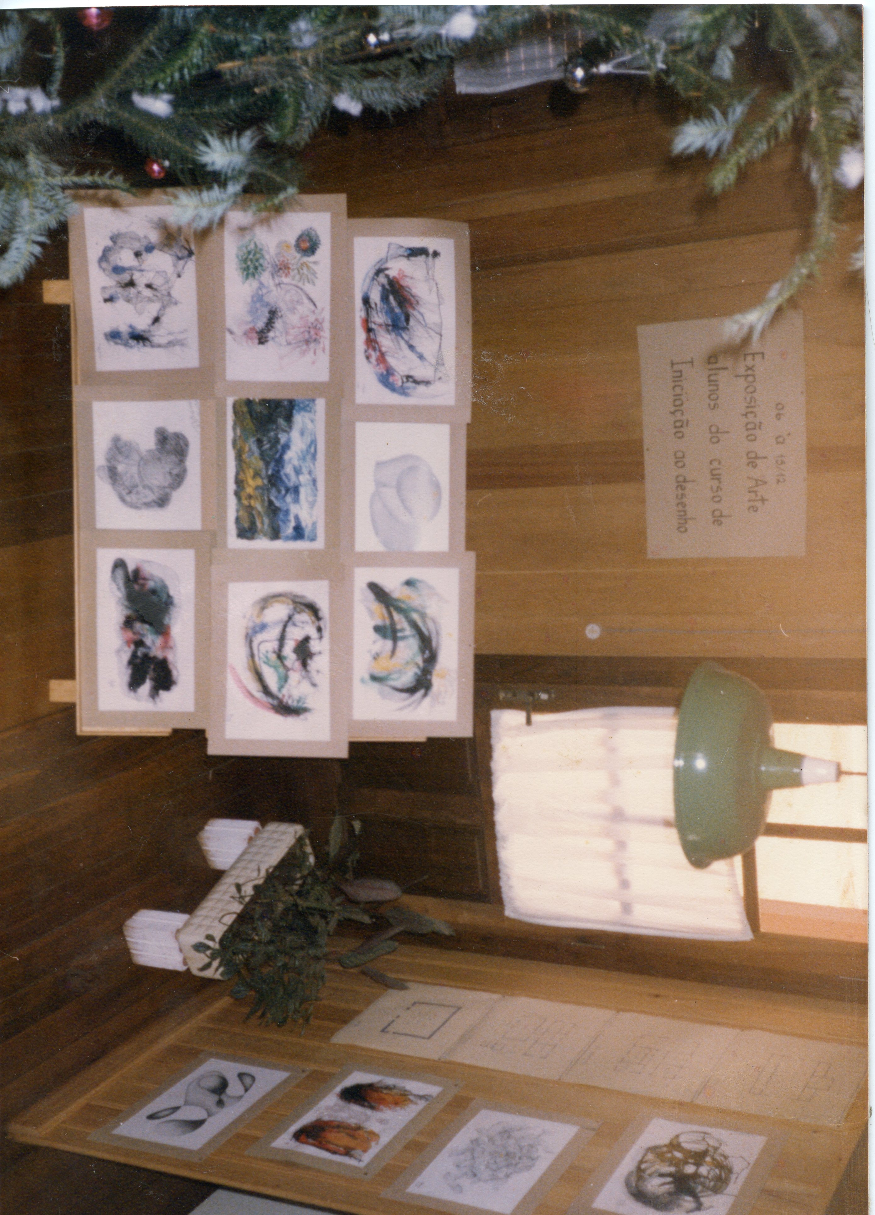 Uma fotografia de uma exposição de arte de alunos de um curso de desenho, montada em um ambiente com paredes de madeira. O espaço é decorado com elementos natalinos, como um galho de pinheiro, e exibe diversos desenhos em papel fixados em suportes, acompanhados por um cartaz informativo.