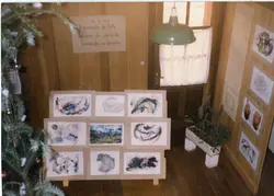 Uma fotografia de uma exposição de arte de alunos de um curso de desenho, montada em um ambiente com paredes de madeira. O espaço é decorado com elementos natalinos, como um galho de pinheiro, e exibe diversos desenhos em papel fixados em suportes, acompanhados por um cartaz informativo.