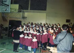 Uma fotografia de um coral infantil apresentando-se em um ginásio de esportes, com uma regente à esquerda e um músico com violão à direita. O cartaz na parede menciona 'Cresce Videira! Administração 93/96', situando o evento em Videira, Santa Catarina, durante a década de 1990.