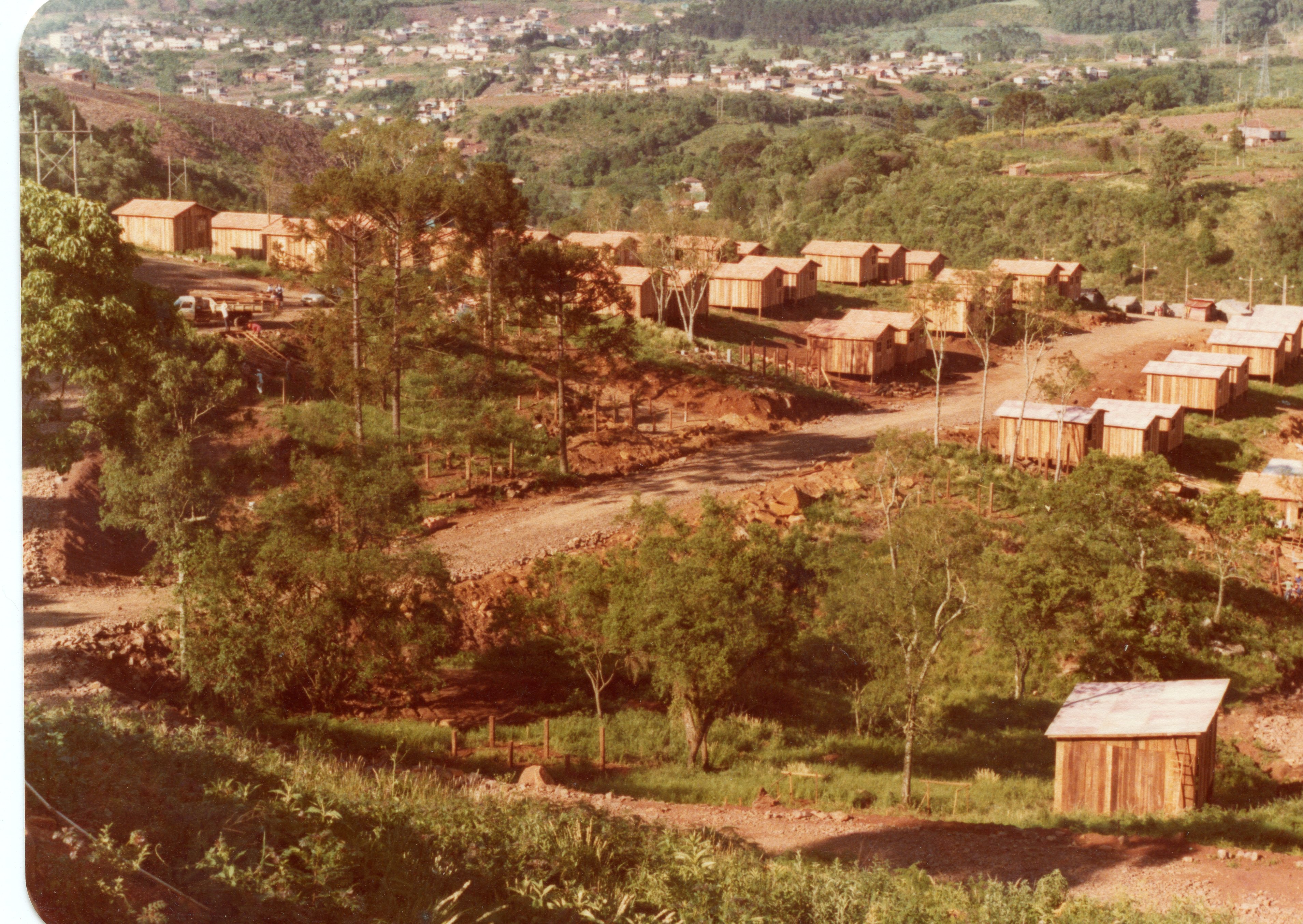 Uma vista aérea de um assentamento ou loteamento em fase inicial de construção, caracterizado por várias casas de madeira simples dispostas em um terreno montanhoso. A paisagem sugere uma região de colonização no interior do Brasil, possivelmente nas décadas de 1970 ou 1980, com vegetação nativa e estradas de terra ainda em formação.