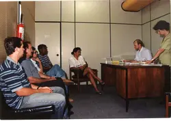 Uma fotografia interna que mostra um grupo de pessoas aguardando atendimento em um escritório ou repartição pública. O ambiente possui divisórias de escritório típicas das décadas de 1980 ou 1990, com pessoas sentadas em cadeiras e uma mesa de atendimento ao fundo.