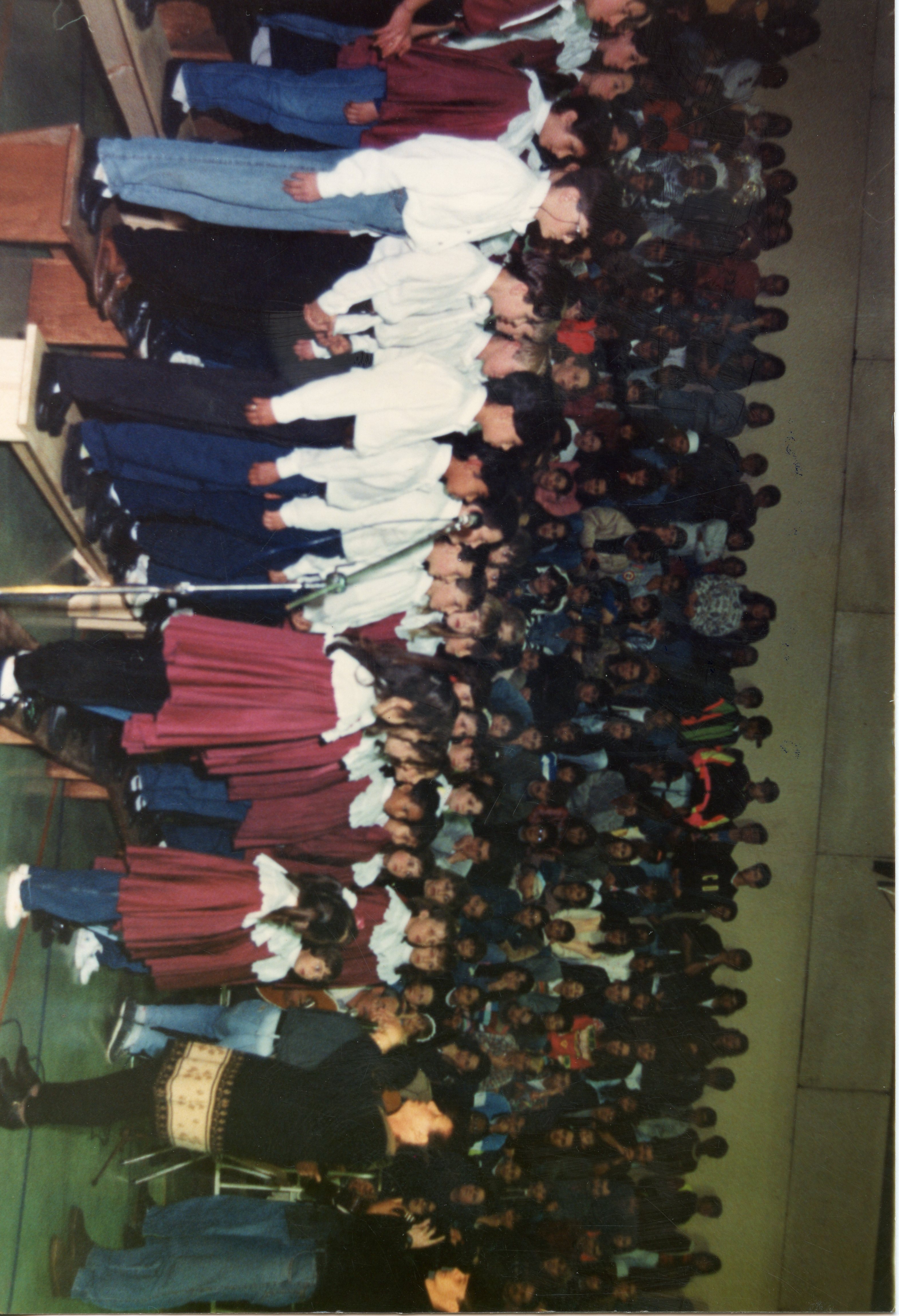 Uma fotografia colorida que registra um evento escolar ou comunitário, possivelmente uma apresentação de coral ou formatura, com um grande grupo de pessoas reunidas em um espaço interno. As vestimentas, incluindo camisas brancas e saias vermelhas, sugerem um padrão de uniforme escolar típico das décadas de 1980 ou 1990.