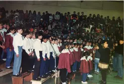 Uma fotografia colorida que registra um evento escolar ou comunitário, possivelmente uma apresentação de coral ou formatura, com um grande grupo de pessoas reunidas em um espaço interno. As vestimentas, incluindo camisas brancas e saias vermelhas, sugerem um padrão de uniforme escolar típico das décadas de 1980 ou 1990.