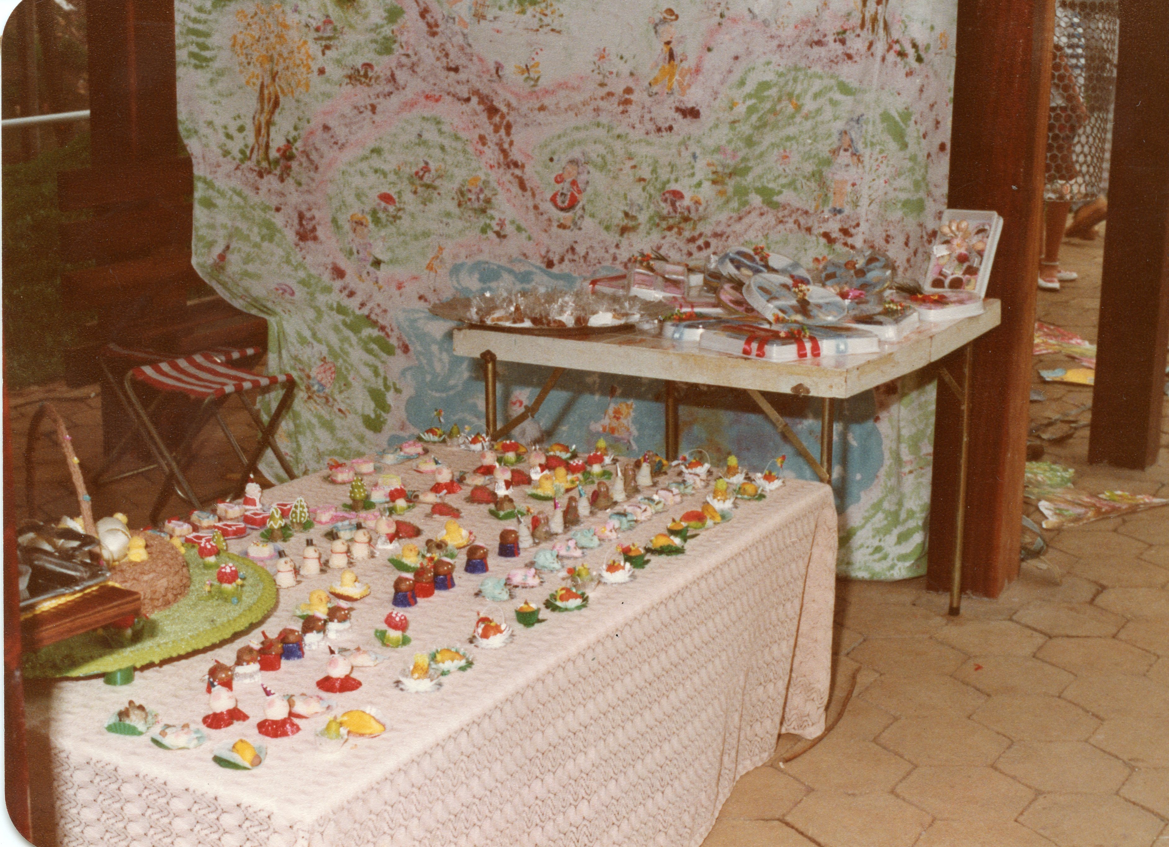 Uma mesa coberta com toalha de renda exibe uma grande variedade de pequenos doces decorados, possivelmente para uma festa infantil ou evento comemorativo. Ao fundo, um painel ilustrado e uma mesa auxiliar com embalagens sugerem um ambiente de celebração caseira ou comunitária, típico das décadas de 1980 ou 1990.