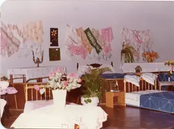 Uma exposição de artesanato doméstico, provavelmente em um centro comunitário ou salão paroquial, exibindo toalhas de mesa bordadas, colchas e trabalhos manuais. O ambiente apresenta móveis de madeira simples e arranjos florais, sugerindo uma feira de artesanato ou exposição cultural típica de pequenas cidades brasileiras nas décadas de 1970 ou 1980.