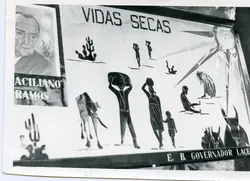 Uma fotografia em preto e branco de um cartaz ou painel escolar ilustrando a obra 'Vidas Secas' de Graciliano Ramos. O painel apresenta desenhos estilizados de uma família retirante, cactos e um sol forte, típicos do cenário da seca no sertão brasileiro, com o retrato do autor no canto superior esquerdo.