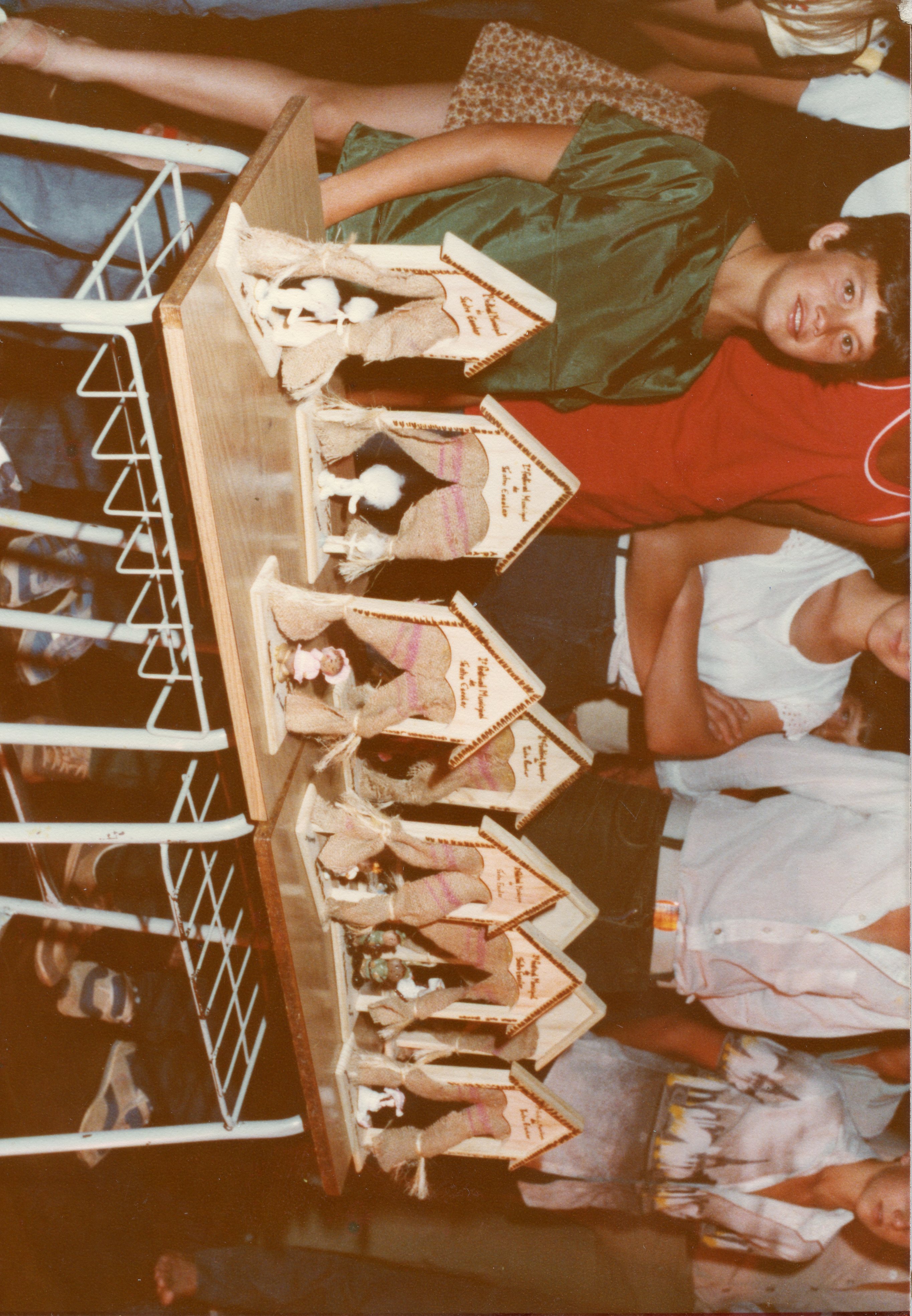 Uma fotografia de ângulo superior mostrando uma mesa com vários troféus ou lembranças artesanais em formato de casinha, decorados com juta. Ao redor da mesa, jovens e crianças estão reunidos, sugerindo um evento escolar ou comunitário, possivelmente em uma cidade pequena brasileira durante a década de 1980 ou 1990.