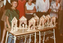 Uma fotografia de ângulo superior mostrando uma mesa com vários troféus ou lembranças artesanais em formato de casinha, decorados com juta. Ao redor da mesa, jovens e crianças estão reunidos, sugerindo um evento escolar ou comunitário, possivelmente em uma cidade pequena brasileira durante a década de 1980 ou 1990.