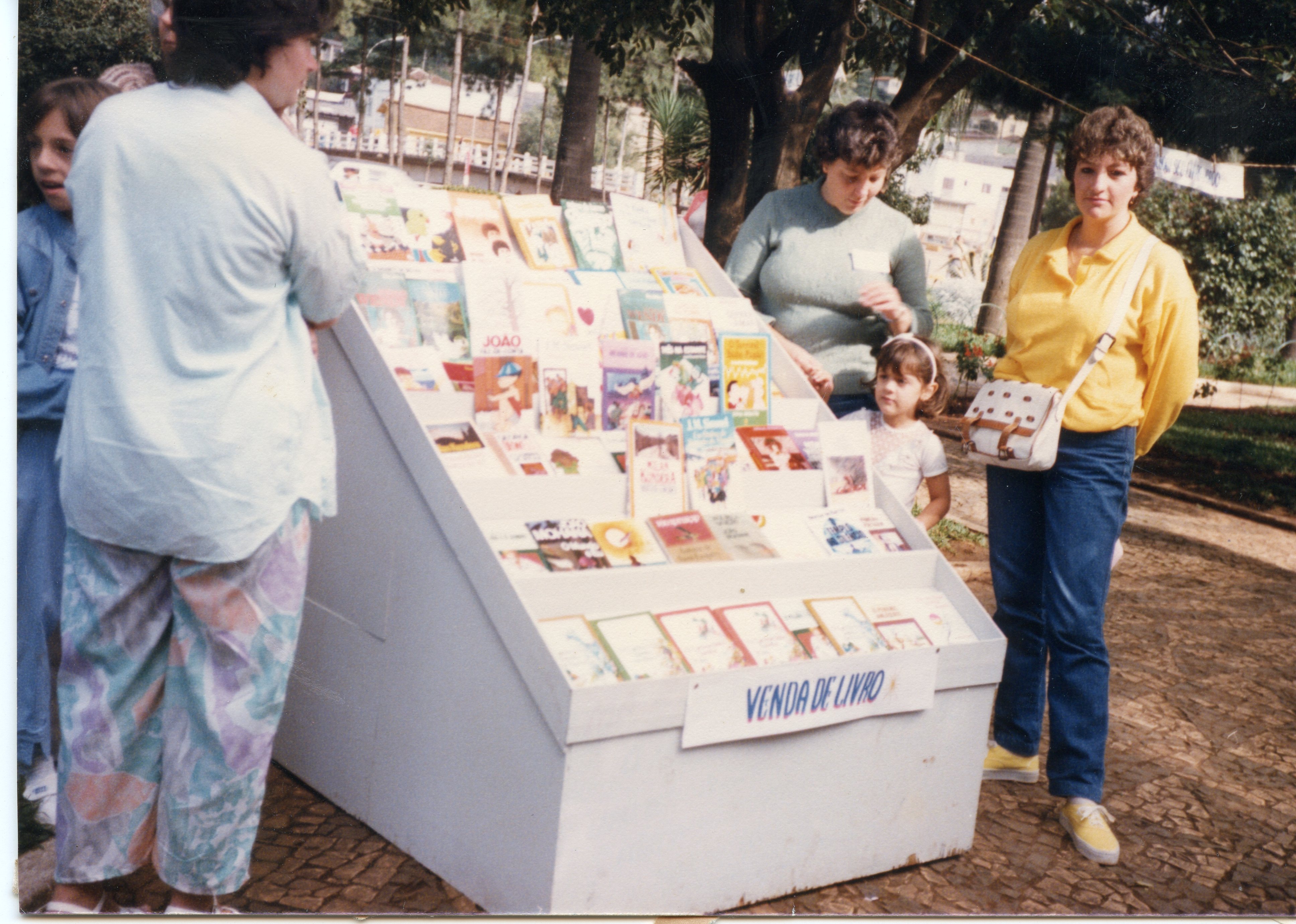 Uma cena ao ar livre mostrando uma pequena feira ou exposição de livros em uma praça pública, com pessoas observando as obras expostas em um suporte de madeira. O estilo das roupas e a qualidade da fotografia colorida sugerem um contexto brasileiro da década de 1980 ou início de 1990.