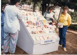 Uma cena ao ar livre mostrando uma pequena feira ou exposição de livros em uma praça pública, com pessoas observando as obras expostas em um suporte de madeira. O estilo das roupas e a qualidade da fotografia colorida sugerem um contexto brasileiro da década de 1980 ou início de 1990.