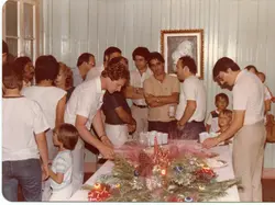 Uma fotografia de uma reunião social ou confraternização familiar em um ambiente interno, com decoração que sugere uma celebração de fim de ano ou natal. Os participantes vestem roupas típicas da década de 1980, com homens de camisas polo e calças de sarja, em um espaço com paredes de madeira pintada.