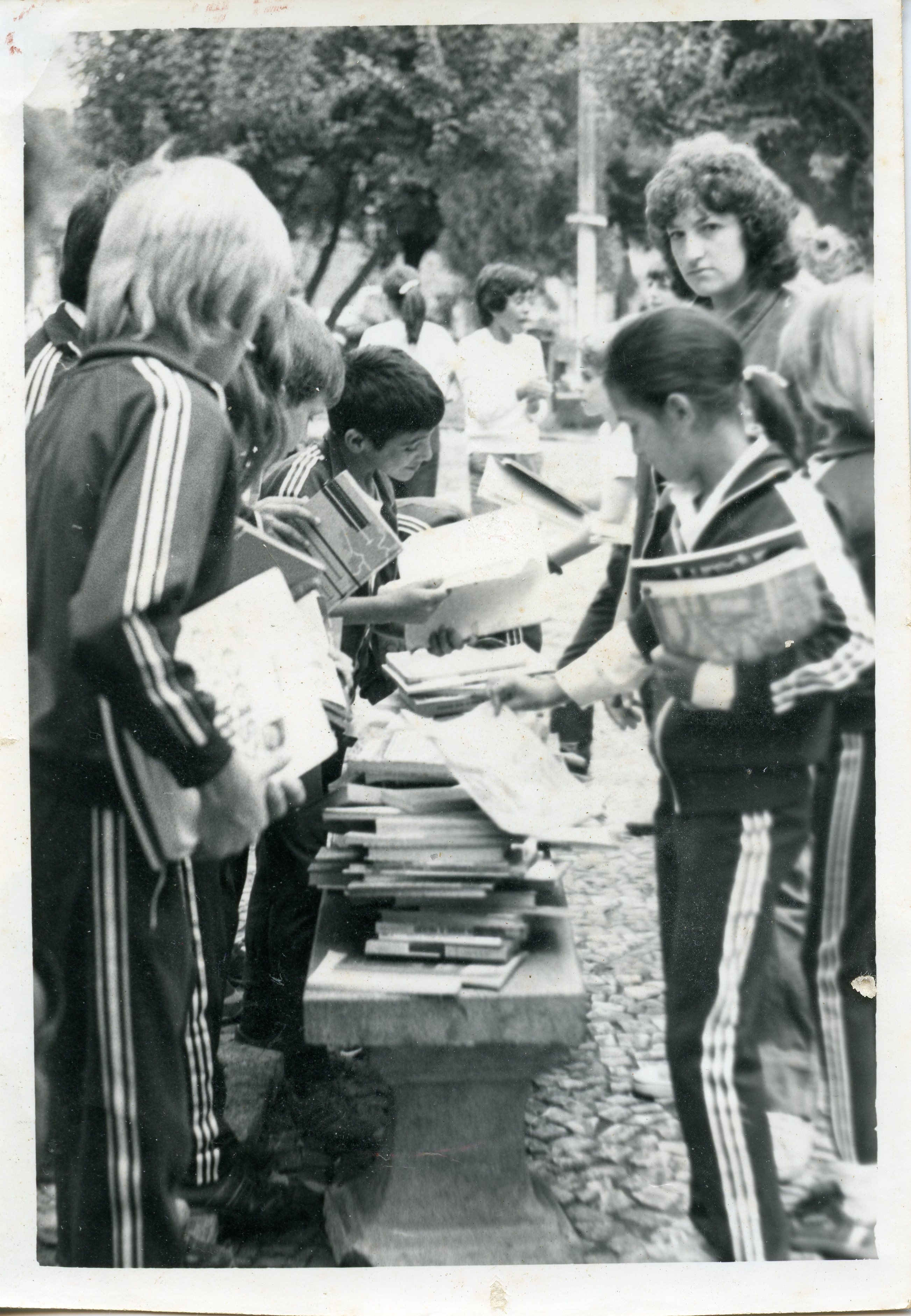Uma fotografia em preto e branco que retrata um grupo de crianças e adolescentes uniformizados com agasalhos esportivos, possivelmente em uma atividade escolar ou feira de livros ao ar livre. O cenário sugere um ambiente público, como uma praça, com os jovens reunidos em torno de uma mesa de pedra empilhada com livros, indicando um evento educacional ou cultural típico de meados da década de 1980.
