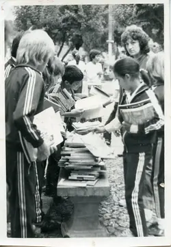 Uma fotografia em preto e branco que retrata um grupo de crianças e adolescentes uniformizados com agasalhos esportivos, possivelmente em uma atividade escolar ou feira de livros ao ar livre. O cenário sugere um ambiente público, como uma praça, com os jovens reunidos em torno de uma mesa de pedra empilhada com livros, indicando um evento educacional ou cultural típico de meados da década de 1980.