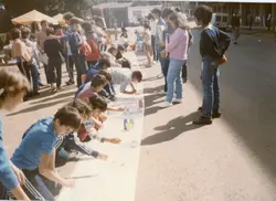 Uma cena ao ar livre mostrando um grupo de jovens e crianças participando de uma atividade de pintura em um longo papel estendido no chão. O estilo das roupas, como agasalhos esportivos e jeans, sugere um contexto escolar ou comunitário típico da década de 1980 ou início de 1990 no Brasil.