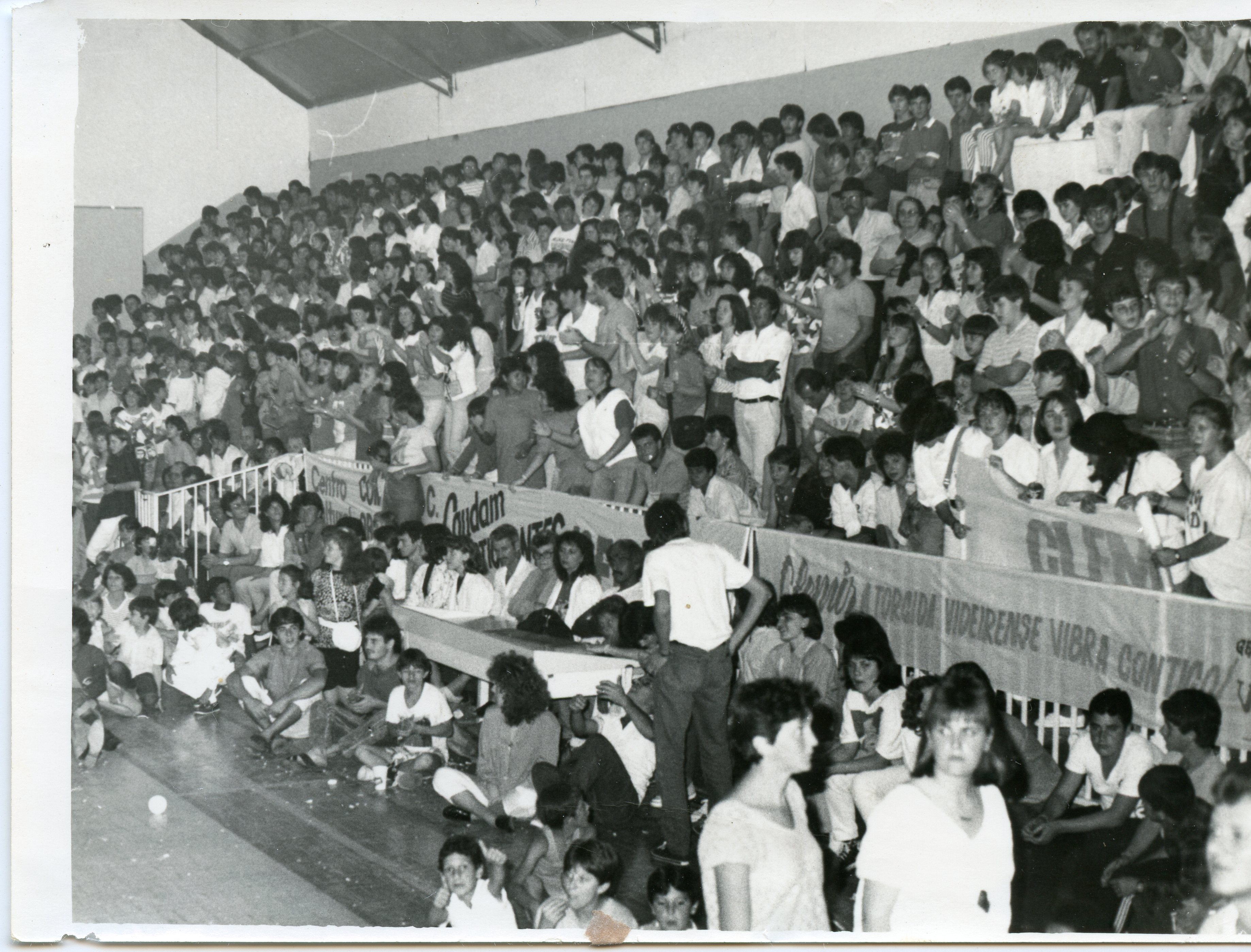 Fotografia em preto e branco registrando uma arquibancada lotada de um ginásio de esportes, provavelmente em Videira, Santa Catarina. O público, composto majoritariamente por jovens, exibe faixas com dizeres como 'A torcida videirense vibra contigo', sugerindo um evento esportivo local ocorrido possivelmente entre as décadas de 1980 e 1990.