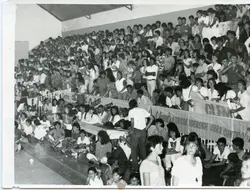 Fotografia em preto e branco registrando uma arquibancada lotada de um ginásio de esportes, provavelmente em Videira, Santa Catarina. O público, composto majoritariamente por jovens, exibe faixas com dizeres como 'A torcida videirense vibra contigo', sugerindo um evento esportivo local ocorrido possivelmente entre as décadas de 1980 e 1990.