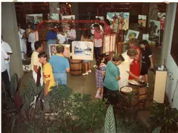 Uma exposição de arte em um ambiente interno, possivelmente um centro cultural ou galeria, com diversas pinturas expostas em estruturas metálicas vermelhas. O público circula pelo espaço, que apresenta elementos rústicos como barris de madeira, sugerindo um evento cultural ou social típico das décadas de 1980 ou 1990.