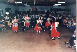 Uma fotografia capturando uma apresentação de dança folclórica, possivelmente de influência italiana, realizada em um ambiente interno com uma grande plateia. Os dançarinos vestem trajes típicos com predominância da cor vermelha, enquanto o público assiste sentado em um espaço que remete a um centro comunitário ou salão de festas típico de colônias no sul do Brasil.