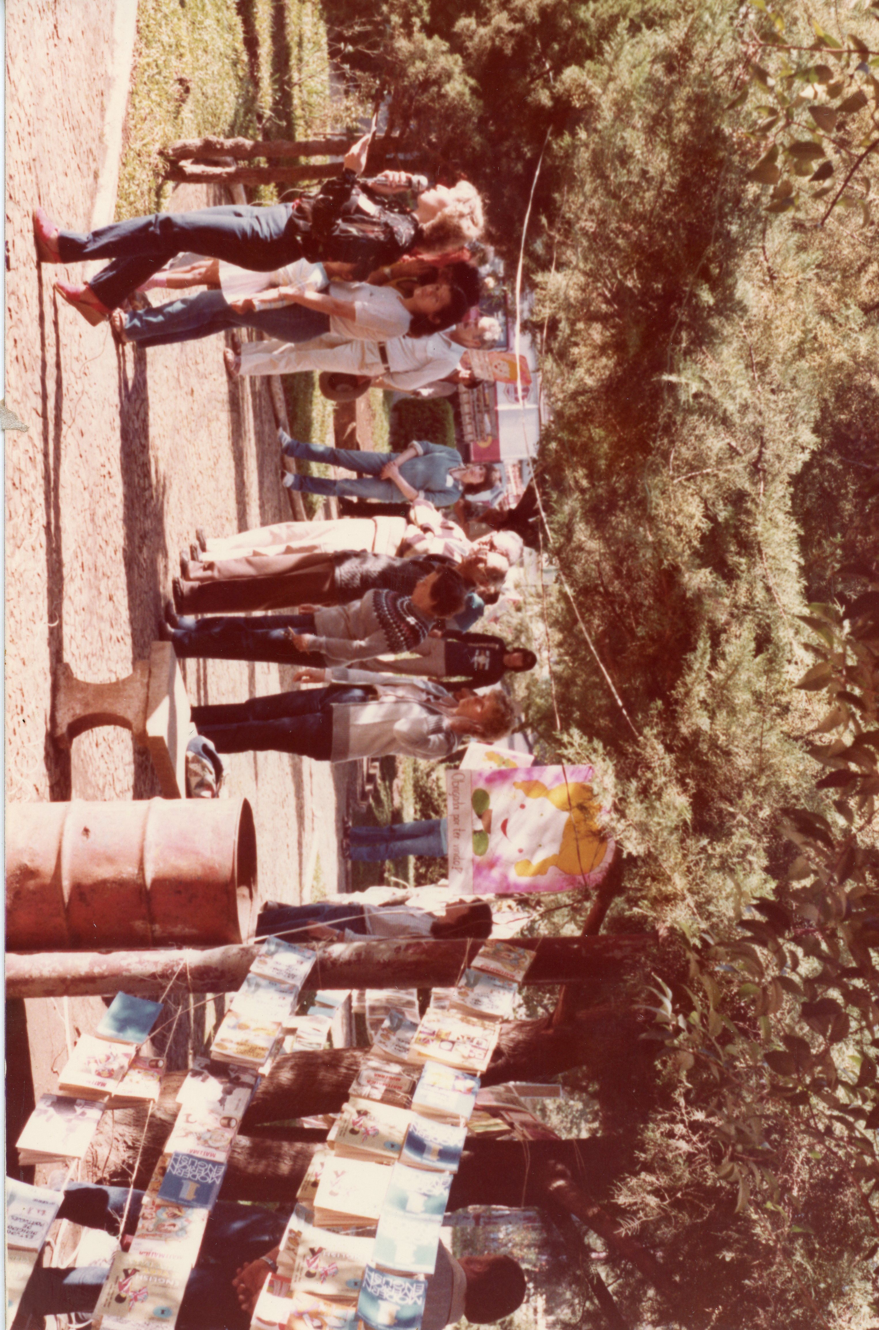 Uma fotografia colorida, possivelmente da década de 1980, mostrando um evento ao ar livre em uma praça ou parque, com pessoas caminhando e interagindo. Há uma exposição de livros ou materiais impressos pendurados em cordas entre árvores, sugerindo uma feira cultural ou escolar, com um cartaz ilustrado visível ao fundo.