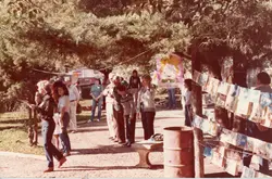 Uma fotografia colorida, possivelmente da década de 1980, mostrando um evento ao ar livre em uma praça ou parque, com pessoas caminhando e interagindo. Há uma exposição de livros ou materiais impressos pendurados em cordas entre árvores, sugerindo uma feira cultural ou escolar, com um cartaz ilustrado visível ao fundo.