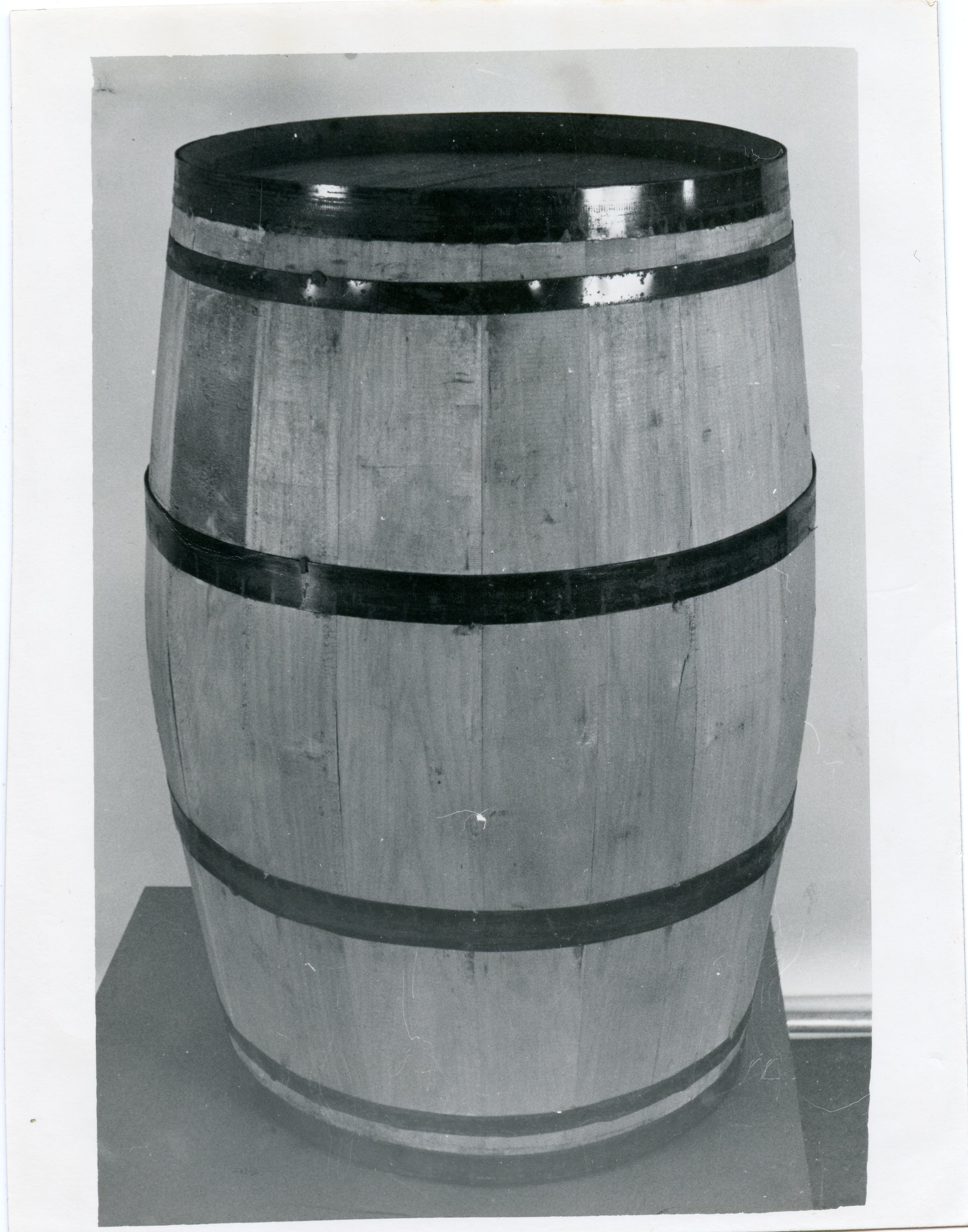 Uma fotografia em preto e branco de um barril de madeira tradicional com aros metálicos, posicionado sobre uma base. Este tipo de objeto era amplamente utilizado na indústria vinícola e no armazenamento de produtos agrícolas, sendo um símbolo comum da história da colonização e da produção de vinhos em regiões como Videira.