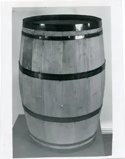 Uma fotografia em preto e branco de um barril de madeira tradicional com aros metálicos, posicionado sobre uma base. Este tipo de objeto era amplamente utilizado na indústria vinícola e no armazenamento de produtos agrícolas, sendo um símbolo comum da história da colonização e da produção de vinhos em regiões como Videira.