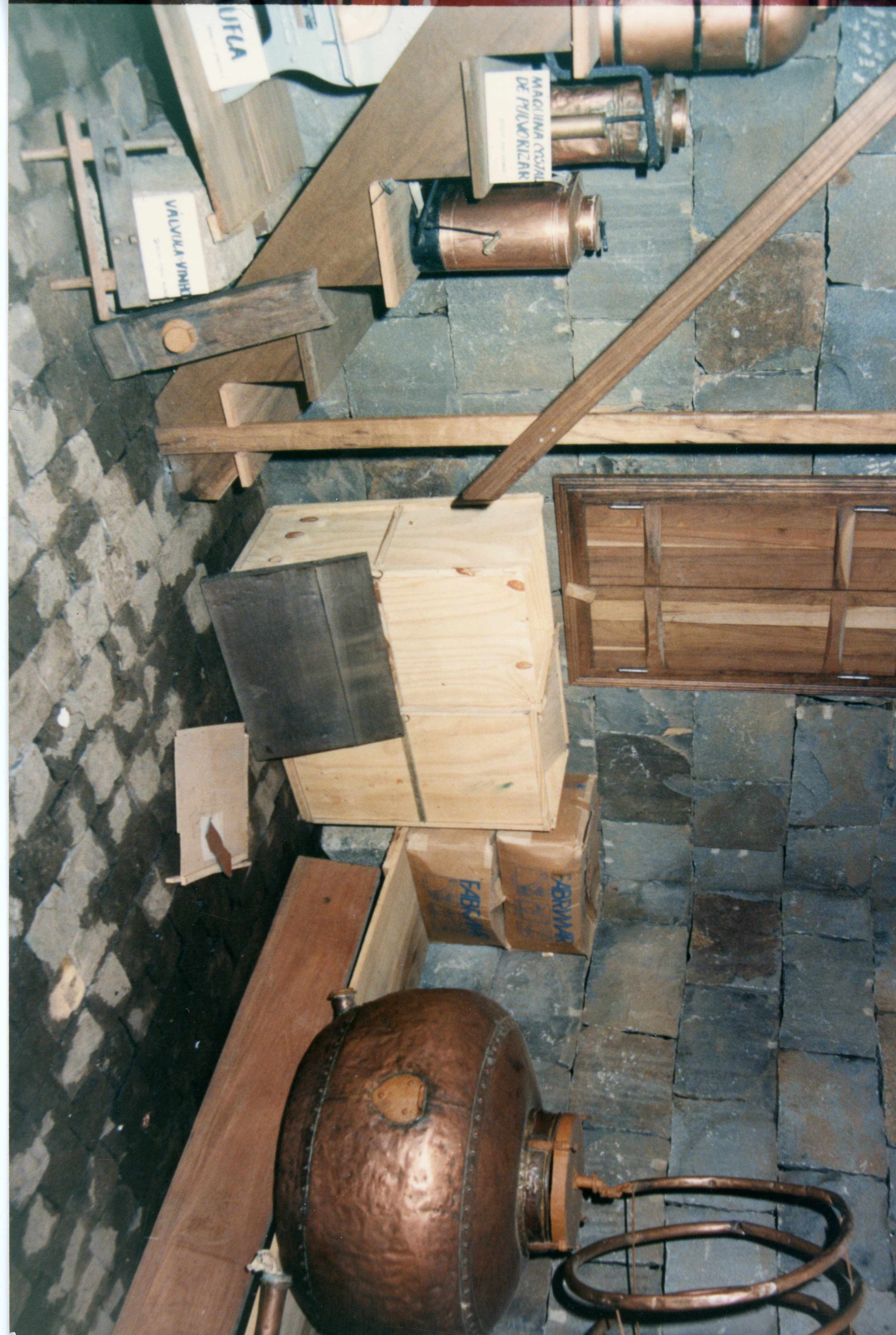 A imagem mostra uma exposição ou acervo de objetos históricos, possivelmente relacionados à agricultura ou produção artesanal, como alambiques de cobre e ferramentas de madeira. O cenário apresenta paredes de pedra rústica e prateleiras improvisadas, sugerindo um museu local ou uma adega antiga, comum em regiões de colonização europeia no sul do Brasil.