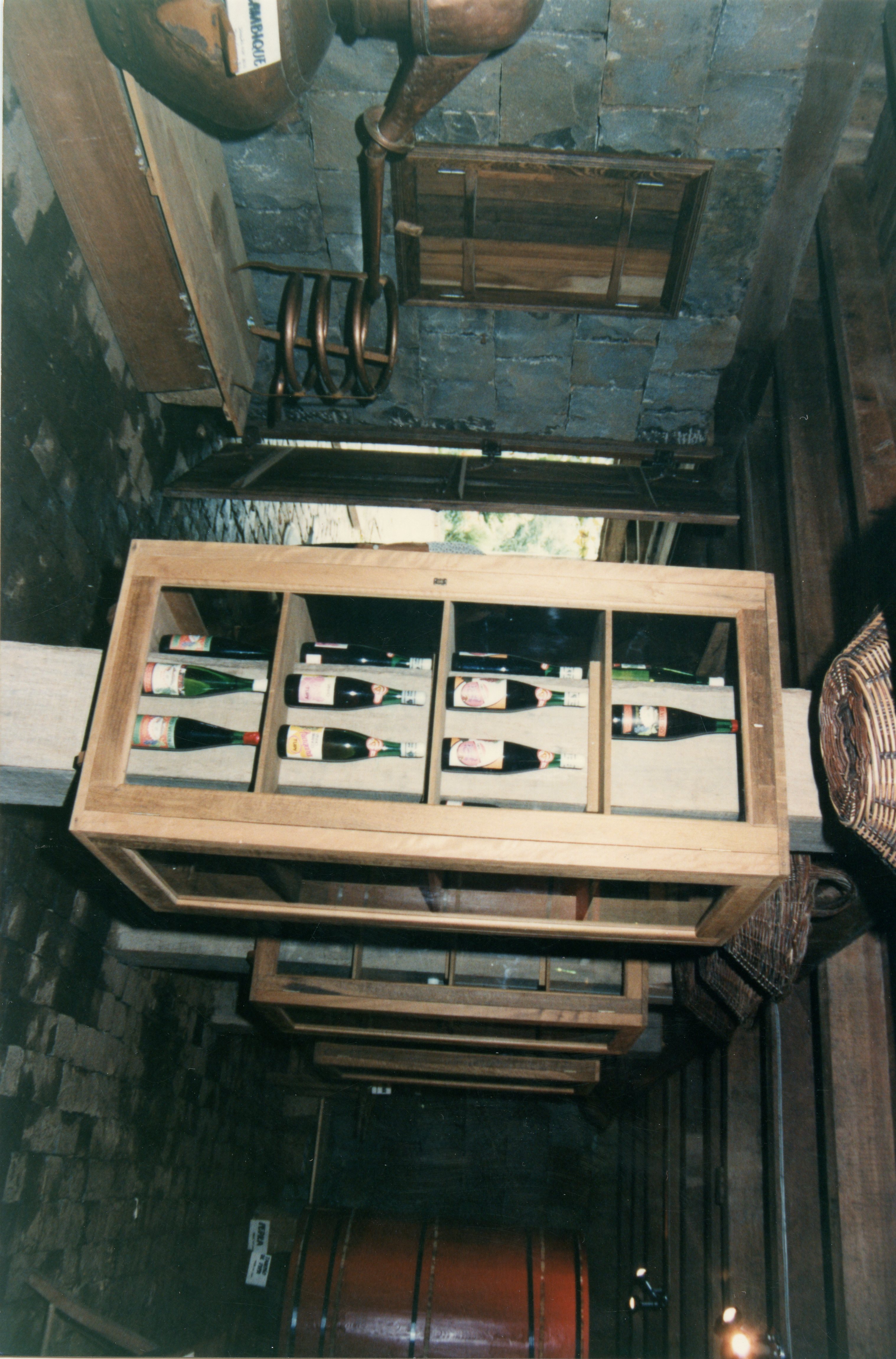 A imagem mostra o interior de uma adega rústica, possivelmente em uma região de colonização italiana como Videira, com garrafas de vinho expostas em prateleiras de madeira. Destacam-se elementos tradicionais como um alambique de cobre no teto e grandes barris de madeira, sugerindo um ambiente de produção artesanal de bebidas.