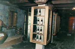 A imagem mostra o interior de uma adega rústica, possivelmente em uma região de colonização italiana como Videira, com garrafas de vinho expostas em prateleiras de madeira. Destacam-se elementos tradicionais como um alambique de cobre no teto e grandes barris de madeira, sugerindo um ambiente de produção artesanal de bebidas.
