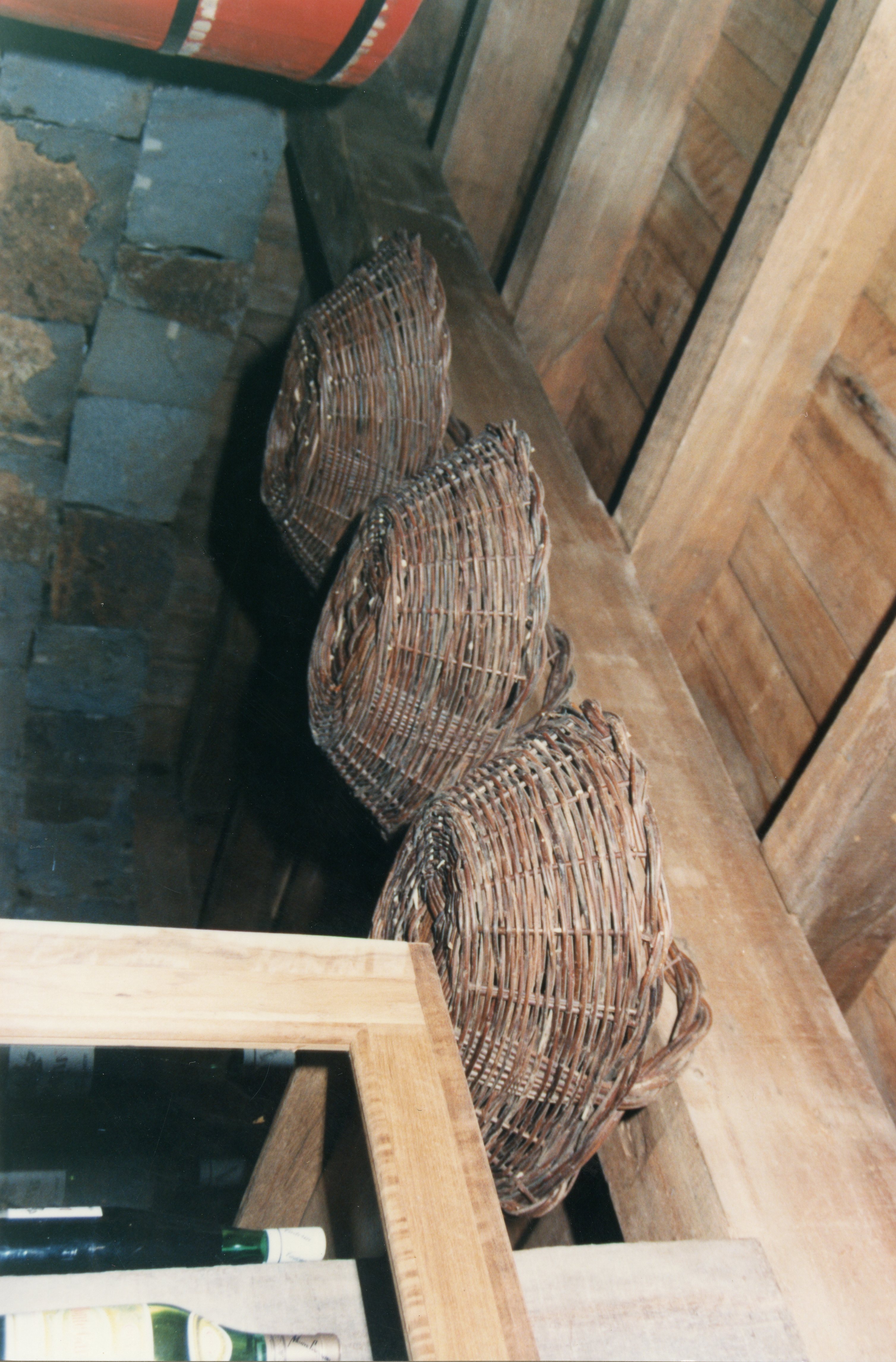 A imagem mostra cestos de vime artesanais pendurados em uma estrutura de madeira rústica, possivelmente em uma adega ou celeiro. O ambiente sugere uma prática agrícola ou de armazenamento tradicional, comum em regiões de colonização europeia no Brasil, como Videira.