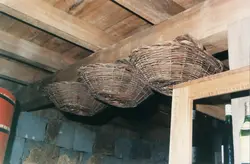 A imagem mostra cestos de vime artesanais pendurados em uma estrutura de madeira rústica, possivelmente em uma adega ou celeiro. O ambiente sugere uma prática agrícola ou de armazenamento tradicional, comum em regiões de colonização europeia no Brasil, como Videira.