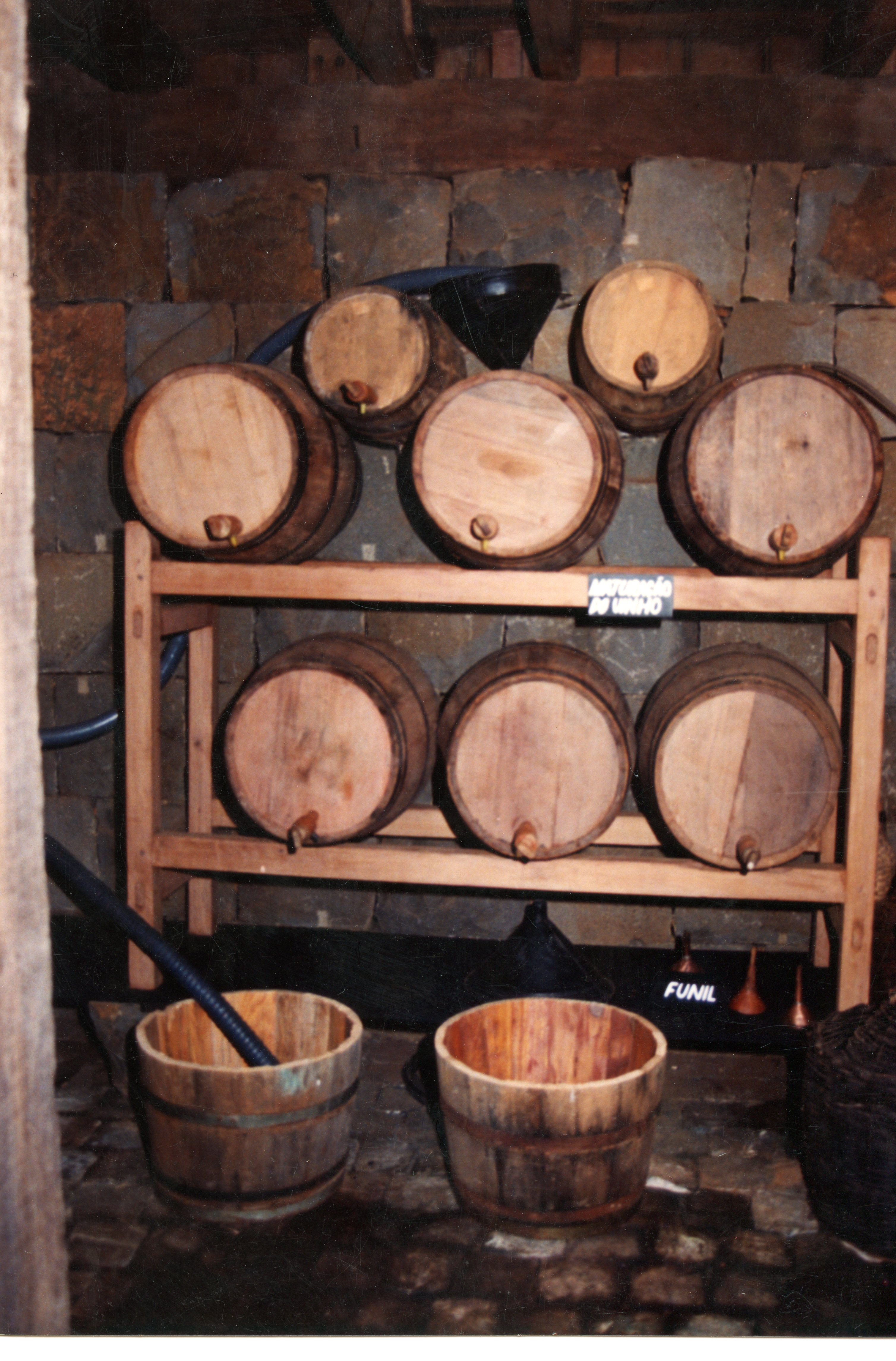 A imagem mostra uma adega rústica com barris de madeira organizados em prateleiras, típicos da produção artesanal de vinho em regiões de colonização italiana, como Videira. O ambiente apresenta paredes de pedra e chão de paralelepípedos, sugerindo um contexto histórico de produção familiar ou pequena indústria vinícola.