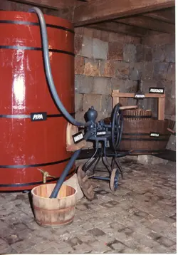 A imagem exibe equipamentos tradicionais de vinificação em um ambiente interno, possivelmente uma adega ou museu em uma região de colonização italiana como Videira. Destacam-se uma grande pipa de vinho vermelha, uma prensa de madeira e uma bomba de remontagem manual, com placas explicativas indicando o uso de cada item.