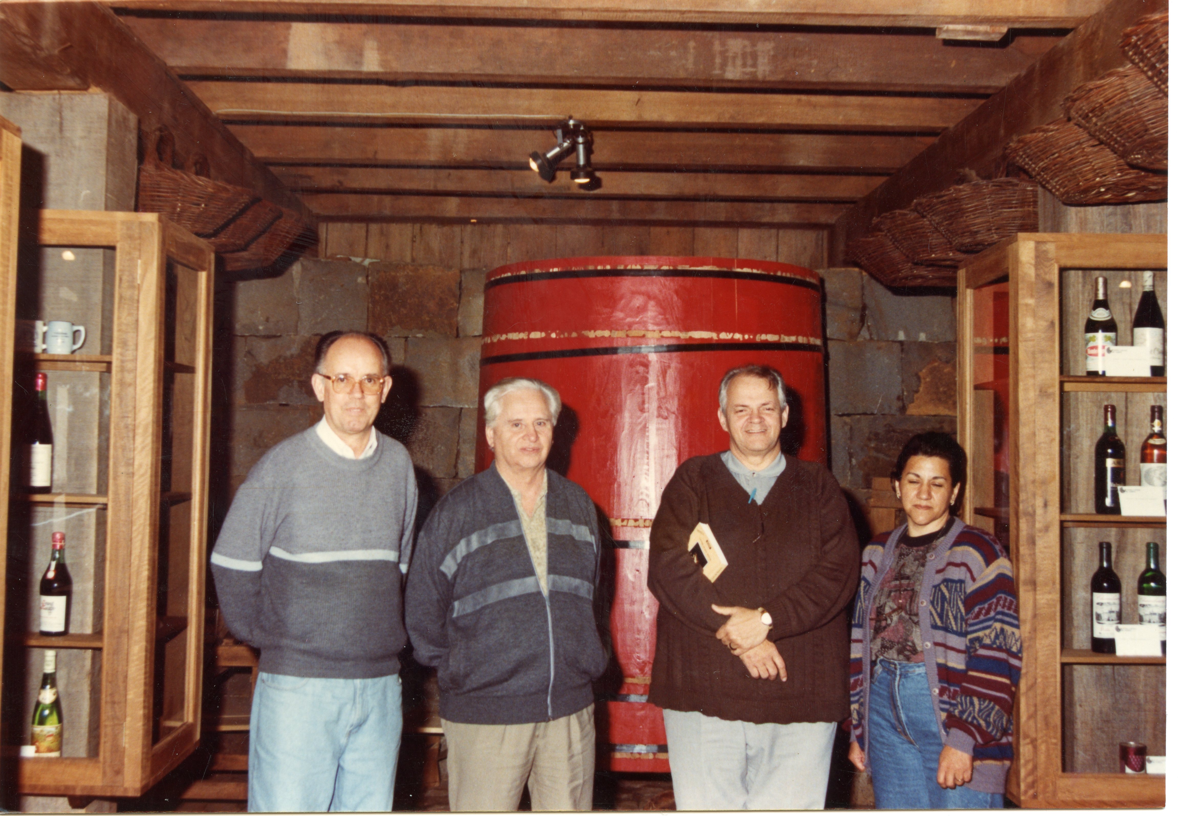Uma fotografia colorida mostrando quatro pessoas posando em uma adega rústica, com um grande barril de vinho vermelho ao fundo. O ambiente apresenta prateleiras de madeira com garrafas de vinho e vigas expostas no teto, sugerindo uma visita a uma vinícola, possivelmente na região de Videira, Santa Catarina. A vestimenta dos indivíduos indica um estilo casual típico do final do século XX.
