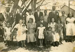 Fotografia de estúdio ou retrato de família numerosa posada ao ar livre, possivelmente em uma área rural ou periurbana. Os adultos vestem trajes formais típicos da década de 1950, como ternos e vestidos, sugerindo um evento comemorativo ou um registro familiar importante.