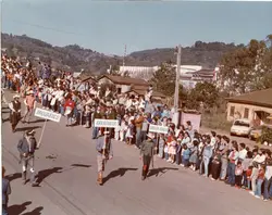 Uma fotografia colorida que registra um desfile cívico ou comunitário em uma pequena cidade brasileira, possivelmente na região de Videira. Homens vestidos com trajes típicos carregam placas identificando localidades como Taquaruçu, Garaguata e Santa Maria, enquanto uma multidão observa o evento ao longo de uma rua de terra batida.