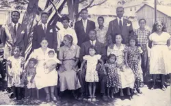 Uma fotografia de grupo de uma família extensa, composta por homens de terno, mulheres com vestidos formais e várias crianças, posando ao ar livre em um ambiente rural ou semi-urbano. A imagem, provavelmente da década de 1950, reflete um retrato de família tradicional com vestimentas típicas da época e uma estrutura de madeira ao fundo.