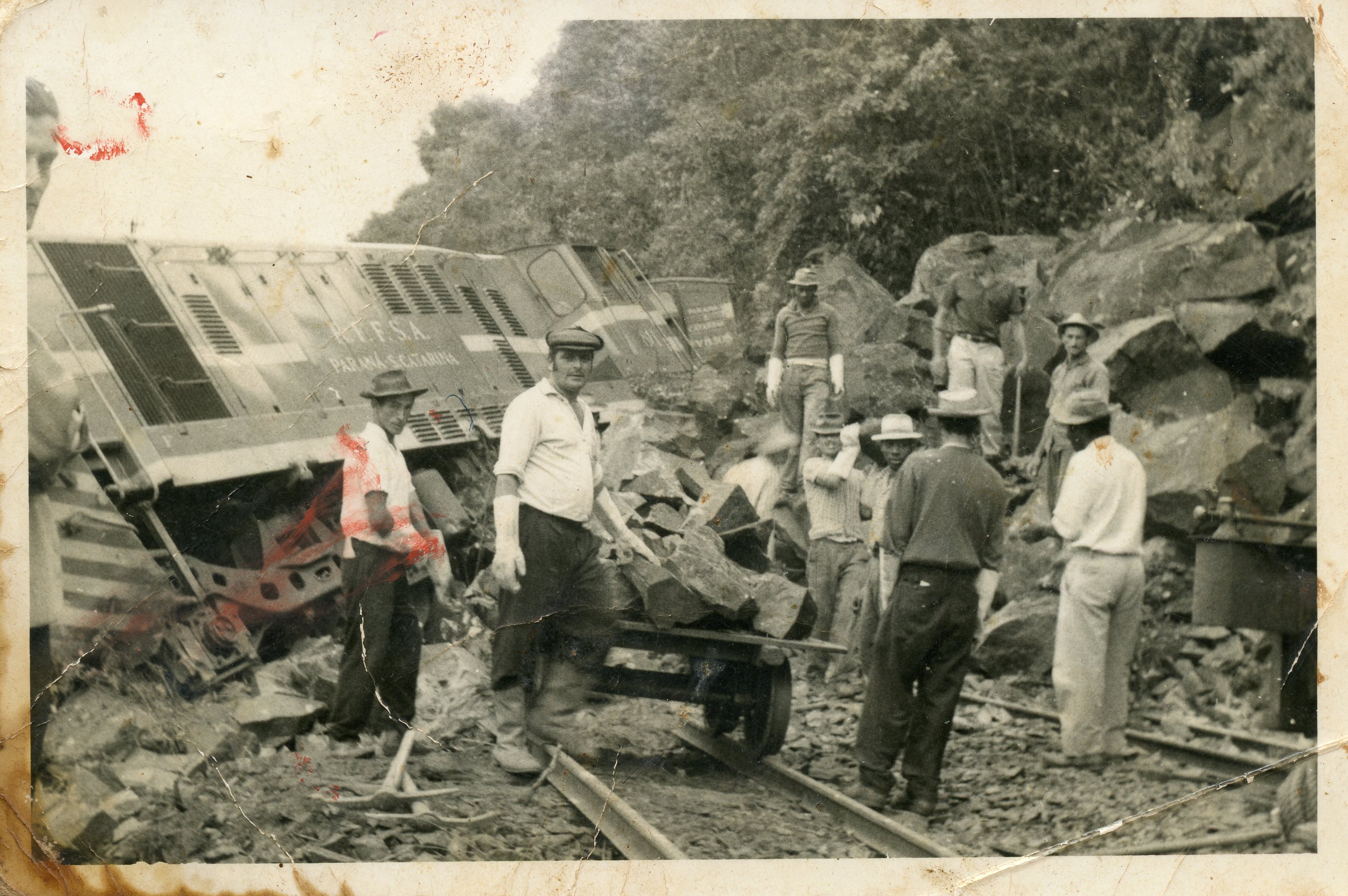 Fotografia histórica em preto e branco mostrando um grupo de trabalhadores ferroviários removendo pedras após um deslizamento ou acidente que atingiu uma locomotiva da RFFSA. A cena, provavelmente situada em uma região montanhosa do sul do Brasil, retrata o esforço manual para liberar os trilhos, com homens vestindo roupas de trabalho típicas de meados do século XX.