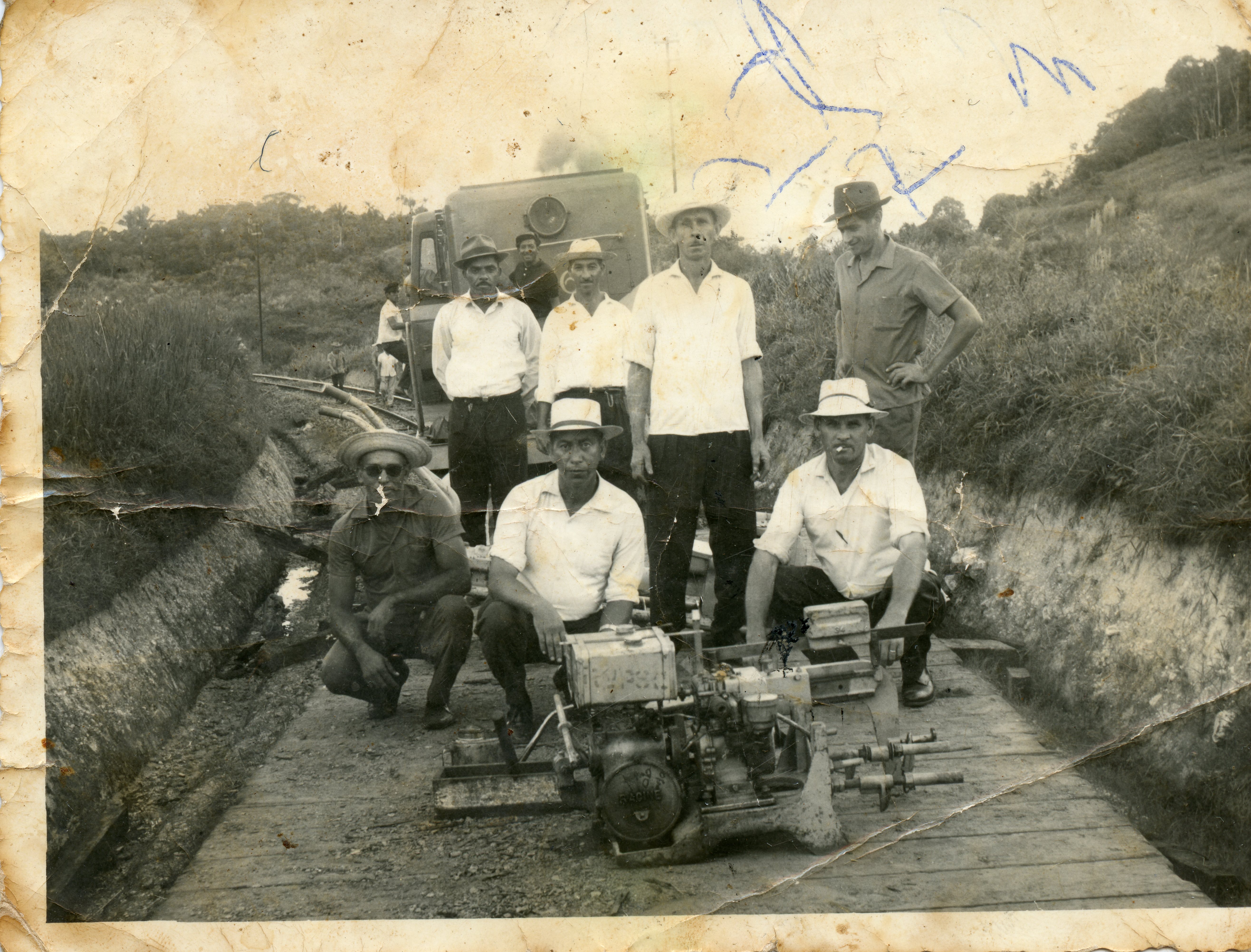 Fotografia em preto e branco retratando um grupo de homens posando ao lado de um equipamento mecânico, possivelmente uma máquina de manutenção ferroviária, em uma área rural. A imagem apresenta desgaste físico significativo, com marcas de dobras e manchas, sugerindo um registro histórico de meados do século XX relacionado a trabalhos de infraestrutura ou colonização.