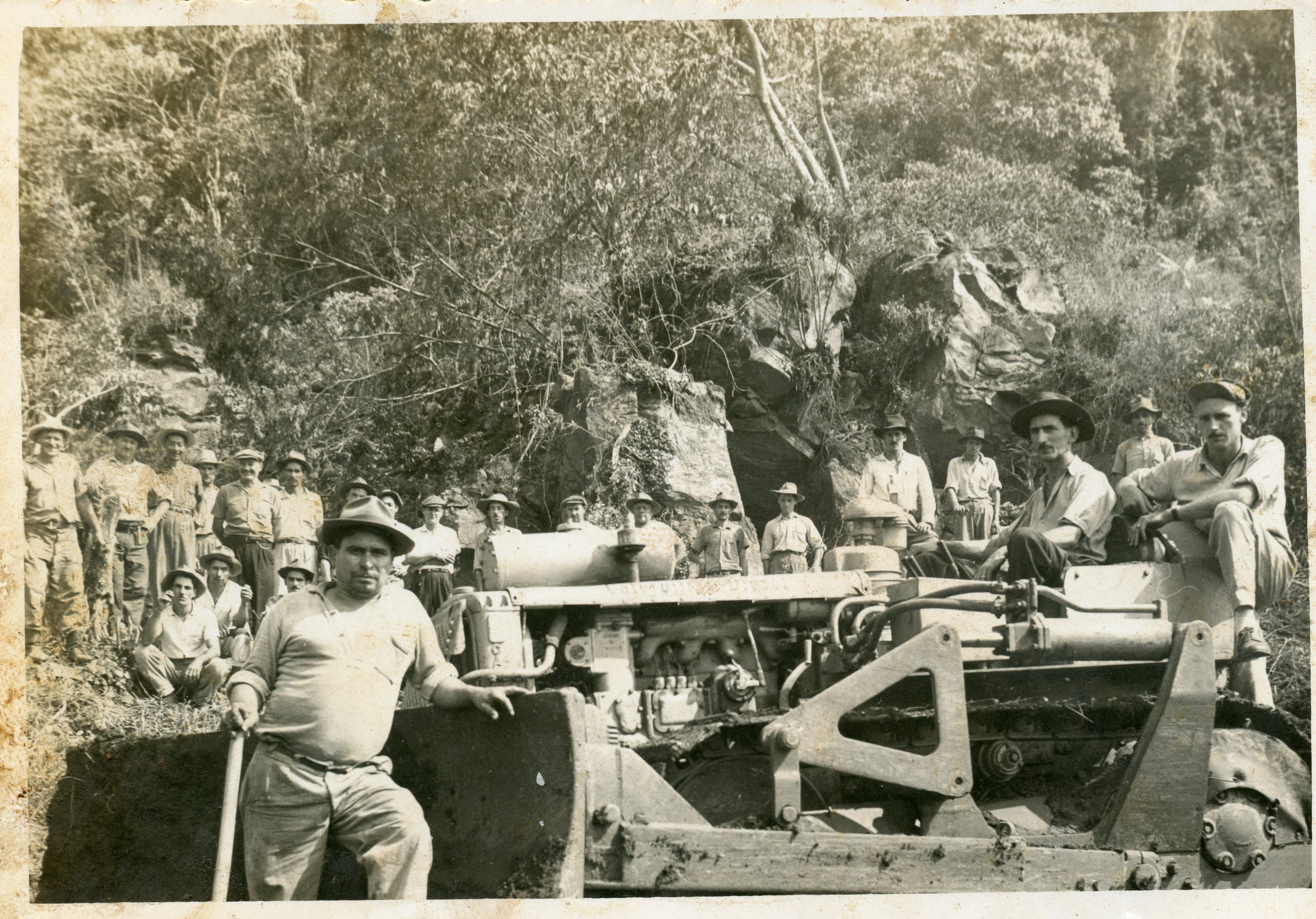 Uma fotografia em preto e branco que retrata um grupo de trabalhadores posando ao lado de um trator de esteira pesado em uma área de desmatamento ou abertura de estradas. A cena sugere um contexto de colonização ou infraestrutura rural no Brasil, possivelmente em meados do século XX, com homens vestindo chapéus e roupas de trabalho simples em um terreno acidentado.