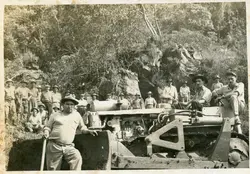 Uma fotografia em preto e branco que retrata um grupo de trabalhadores posando ao lado de um trator de esteira pesado em uma área de desmatamento ou abertura de estradas. A cena sugere um contexto de colonização ou infraestrutura rural no Brasil, possivelmente em meados do século XX, com homens vestindo chapéus e roupas de trabalho simples em um terreno acidentado.
