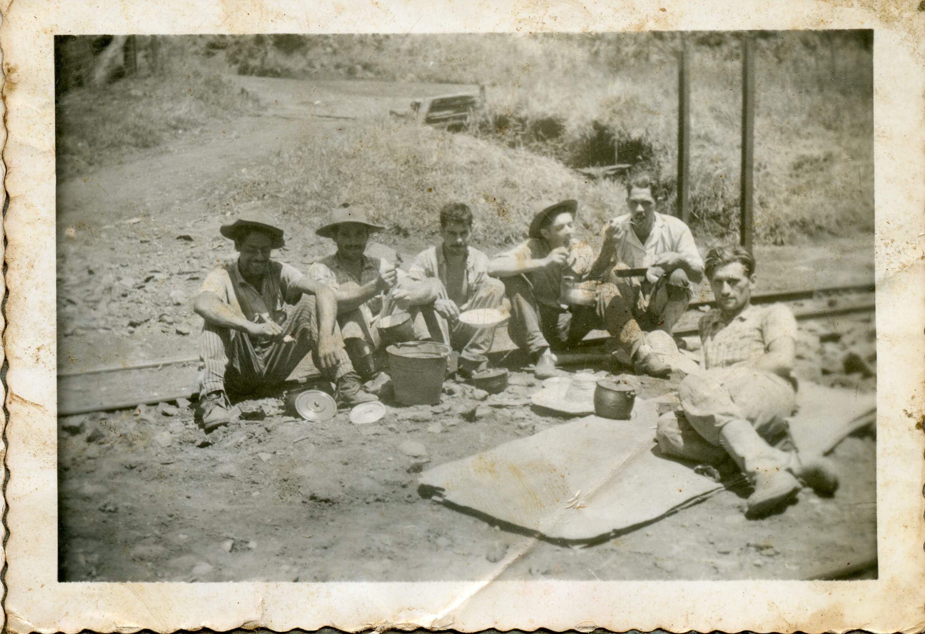 Uma fotografia em preto e branco que retrata um grupo de seis homens fazendo uma refeição ao ar livre, possivelmente trabalhadores ferroviários ou rurais em uma pausa. Eles estão sentados diretamente no solo, próximo a trilhos, utilizando utensílios simples como panelas e pratos, vestindo roupas de trabalho típicas da primeira metade do século XX.